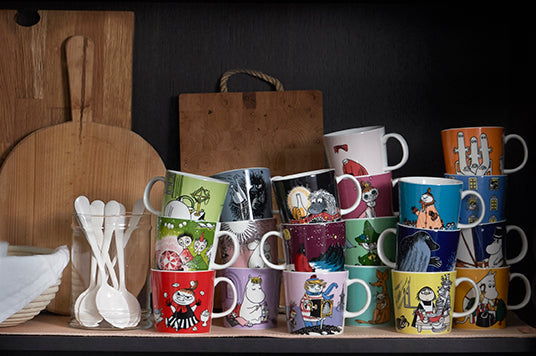 Viele verschiedene Mumin-Becher von Moomin by Arabia mit Szenen aus dem Mumintal in einem Regal